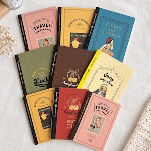 May include: A collection of eight rectangular notebooks in various colors including pink, yellow, blue, olive green, and brown. Each notebook features a black spine, decorative borders, and unique illustrations or text such as Travel Notebook, Pausing for a Moment, Lucky, and Every Tiny Shine.