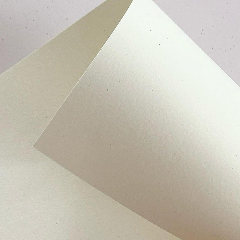 May include: A sheet of off-white, cream-colored paper with a subtle, speckled texture throughout. The paper is folded at an angle, creating a sharp, clean crease that divides the surface into two distinct planes with varying light reflections.