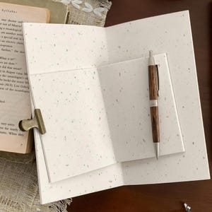 May include: An open notebook with off-white, speckled paper pages and a brass-colored metal binder clip attached to the side. A wooden ballpoint pen with silver-toned metal accents rests on the open pages of the notebook, which is placed on a book.
