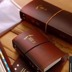 May include: Two dark brown leather-covered notebooks with gold foil text reading Paper Room and Elestore design. Each notebook is secured with a thin orange elastic band and features two silver metal rivets on the spine, holding the cover together.