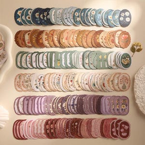 May include: A large collection of oval-shaped paper stickers arranged in six horizontal rows, each featuring various floral illustrations. The stickers are organized by color, including shades of blue, brown, green, cream, purple, and pink.