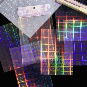 May include: A collection of transparent sheets featuring a holographic grid pattern that reflects rainbow colors. One sheet includes a header card with the text Colorful Light Dream and FLUORESCENT MAGIC. The sheets have a shimmering, iridescent finish.