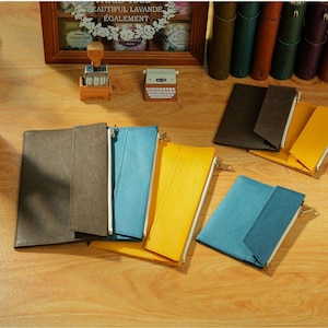 May include: A collection of rectangular zippered pouches in brown, blue, and yellow, featuring a fold-over flap design. The pouches are arranged on a wooden surface alongside a wooden stamp, a miniature typewriter-shaped object, and several upright notebooks.