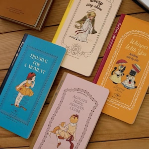 May include: Four rectangular notebooks with illustrated covers. One is blue with text "PAUSING FOR A MOMENT", one is cream with an angel and "Every tiny shine", one is pink with "ALWAYS HERE ALWAYS CLOSE", and one is orange with "Whisper With You".