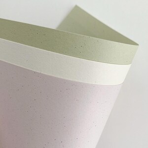 May include: Three stacked sheets of textured paper with a speckled finish. The top sheet is a muted sage green, the middle sheet is a solid off-white, and the bottom sheet is a soft lavender. Each sheet features small dark flecks throughout.