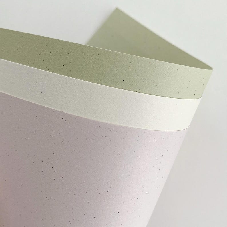 May include: Three stacked sheets of textured paper with a speckled finish. The top sheet is a muted sage green, the middle sheet is a solid off-white, and the bottom sheet is a soft lavender. Each sheet features small dark flecks throughout.