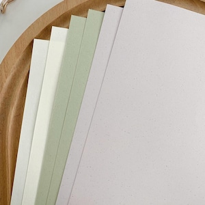 May include: A stack of rectangular paper sheets in varying pastel shades including white, light cream, sage green, and soft lavender. Each sheet features a subtle, speckled texture throughout the surface, arranged in a fan-like overlapping pattern.