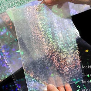 May include: A rectangular transparent plastic sheet featuring a repeating holographic star pattern that reflects rainbow colors. The sheet is held up to display the light-catching finish, with additional sheets visible in the background.