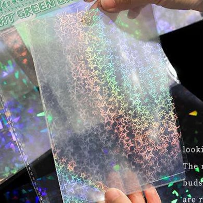 May include: A rectangular transparent plastic sheet featuring a repeating holographic star pattern that reflects rainbow colors. The sheet is held up to display the light-catching finish, with additional sheets visible in the background.