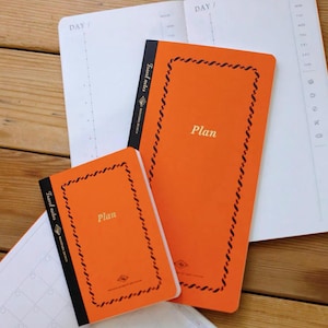 May include: Two orange planners with black spines and a decorative black border. The word Plan is printed in gold on the front covers. The text Travel notes and Reniture Design are visible on the black spines. The planners are open to white pages.