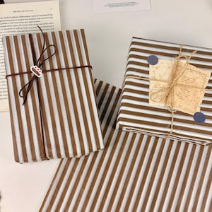 May include: Two rectangular packages wrapped in brown and white striped paper. The left package is tied with a thin brown cord and a small cake charm. The right package is tied with jute twine, featuring a vintage-style paper tag and two blue dots.