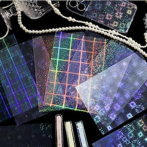 May include: A collection of rectangular transparent sheets featuring holographic patterns including grids, stars, and firework-like bursts. The sheets display a spectrum of iridescent colors against a dark background, accompanied by a string of white pearls.
