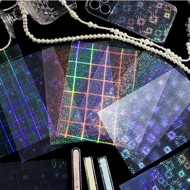 May include: A collection of rectangular transparent sheets featuring holographic patterns including grids, stars, and firework-like bursts. The sheets display a spectrum of iridescent colors against a dark background, accompanied by a string of white pearls.