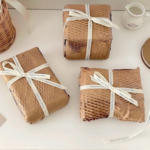 Può includere: Tre pacchetti rettangolari avvolti in carta marrone con trama a nido d'ape, ciascuno legato con un nastro bianco che riporta il testo nero ripetuto "Happy everyday". I pacchetti sono disposti su una superficie piana.