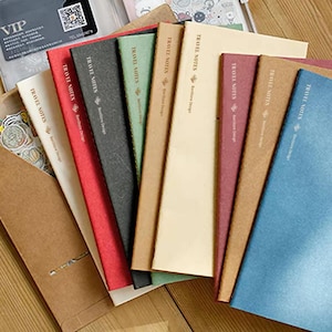 Può includere: Una collezione di quaderni sottili e rettangolari in vari colori, tra cui crema, rosso, nero, verde, marrone chiaro, bordeaux e blu. Ogni copertina presenta il testo TRAVEL NOTES e BenStore Design stampato in orientamento verticale.
