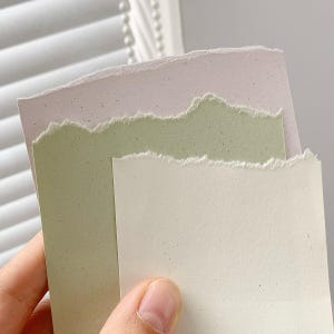 May include: Three rectangular pieces of thick paper with rough, torn edges. The top sheet is a pale lavender, the middle sheet is a soft sage green, and the front sheet is an off-white cream color. Each piece of paper has a subtle speckled texture.