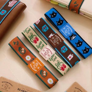 May include: Four rectangular elastic bands featuring woven designs. One is orange with white daisies, one is cream with red cherries and the text Sweet Time, one is brown with blue drink icons, and one is blue with black cat faces. Each has a small label.