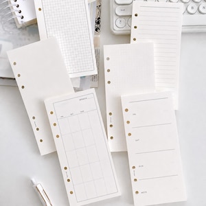 May include: A collection of off-white planner refill pages featuring various layouts including grid, lined, dotted, monthly calendar, and weekly schedule formats. Each rectangular paper sheet has six circular punch holes along the left side.
