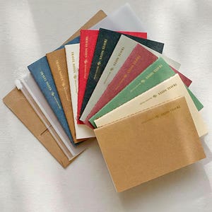 Könnte beinhalten: Eine Sammlung rechteckiger Notizbücher, die fächerförmig angeordnet sind und verschiedene einfarbige Töne wie Blau, Braun, Creme, Rot, Schwarz, Grau, Pink und Grün aufweisen. Jeder Einband ist mit dem Text TRAVEL NOTES und einem Symbol bedruckt.