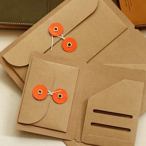 May include: A light brown paper folder featuring a flap secured by two bright orange circular discs and white string. The interior includes a small attached envelope with a matching orange disc closure and three horizontal slots for card storage.