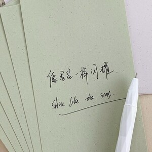 May include: A stack of light sage green rectangular cards with a speckled texture. The top card features black handwritten Chinese characters and the English text "Shine like the stars" with a horizontal line underneath. A white faceted pen rests nearby.