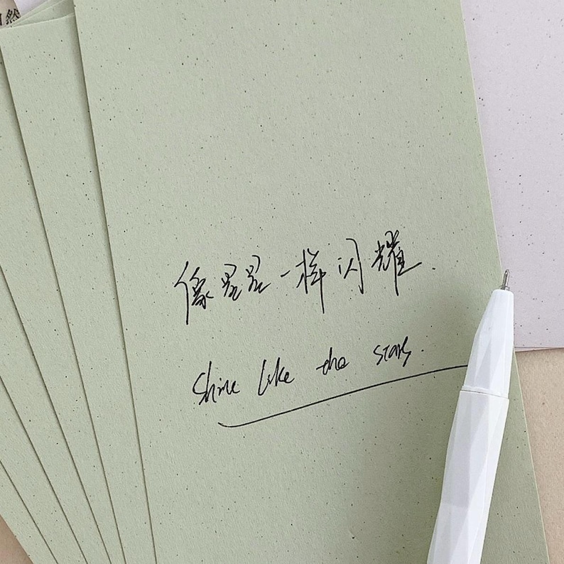 May include: A stack of light sage green rectangular cards with a speckled texture. The top card features black handwritten Chinese characters and the English text "Shine like the stars" with a horizontal line underneath. A white faceted pen rests nearby.