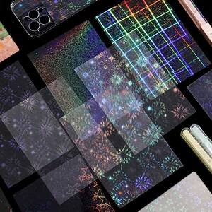 May include: A collection of holographic adhesive sheets and stickers featuring various patterns including fireworks, stars, and grid lines. The designs display a spectrum of iridescent colors that shift against a dark background, alongside a phone case.