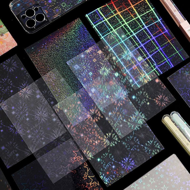 May include: A collection of holographic adhesive sheets and stickers featuring various patterns including fireworks, stars, and grid lines. The designs display a spectrum of iridescent colors that shift against a dark background, alongside a phone case.