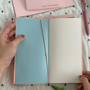 May include: A pink leather-style notebook cover holding an open journal with a light blue left page and a cream-colored right page, secured by a thin pink elastic band. A matching pink notepad labeled TRAVEL NOTES and a pink pen are nearby.
