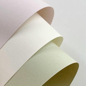May include: Three sheets of thick, textured paper are layered and curled, showcasing a soft color palette. The top sheet is a pale pastel pink, the middle is a creamy off-white, and the bottom sheet is a muted sage green, all with dark specks.