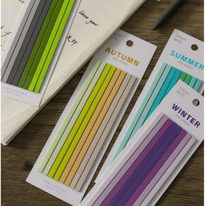 May include: Four sets of thin, rectangular sticky notes on white backing cards. The sets feature color gradients labeled Autumn in gold, Summer in teal, and Winter in purple. Each card contains 200 sheets measuring 140mm x 5mm.