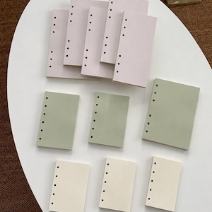 May include: A collection of rectangular paper inserts for a binder, featuring a vertical row of six punched holes along one side. The set includes several pale pink, sage green, and cream-colored sheets arranged in stacks on a white surface.