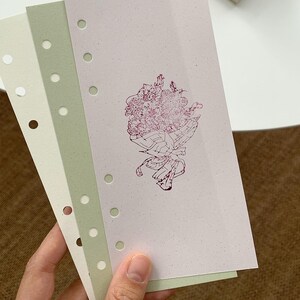 May include: Three rectangular planner inserts with punched holes along the left edge. The sheets are cream, sage green, and light pink. The pink sheet features a dark red line drawing of a floral bouquet tied with a ribbon in the center.