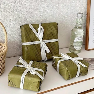 Può includere: Tre pacchetti quadrati avvolti in carta color verde oliva con trama a nido d'ape, ciascuno legato con un nastro bianco che riporta il testo ripetitivo "cleveyer". Una bottiglia di vetro trasparente di bevanda Fentimans Elderflower è accanto.
