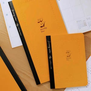 May include: Two orange planners with black spines and the text 2027 Monthly Plan printed on the front covers. The planners feature a decorative border and the words Journey Notes. The planners are displayed on top of open calendar pages.