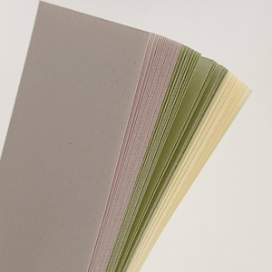 May include: A stack of rectangular paper sheets in a gradient of colors, starting with a muted grey-purple, transitioning through shades of sage green, and ending with a pale cream yellow. Each sheet has a subtle speckled texture throughout.