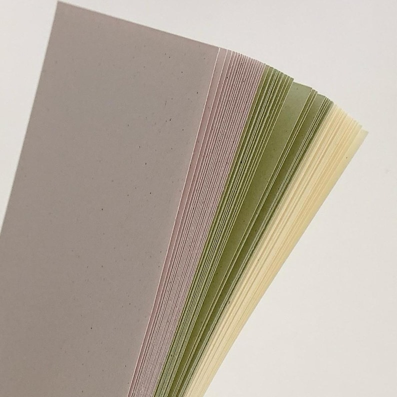 May include: A stack of rectangular paper sheets in a gradient of colors, starting with a muted grey-purple, transitioning through shades of sage green, and ending with a pale cream yellow. Each sheet has a subtle speckled texture throughout.