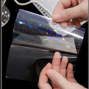 May include: A rectangular sheet of transparent adhesive film featuring a holographic finish that reflects rainbow colors. The film is partially peeled away from its solid black backing, revealing the reflective surface and the clear material.