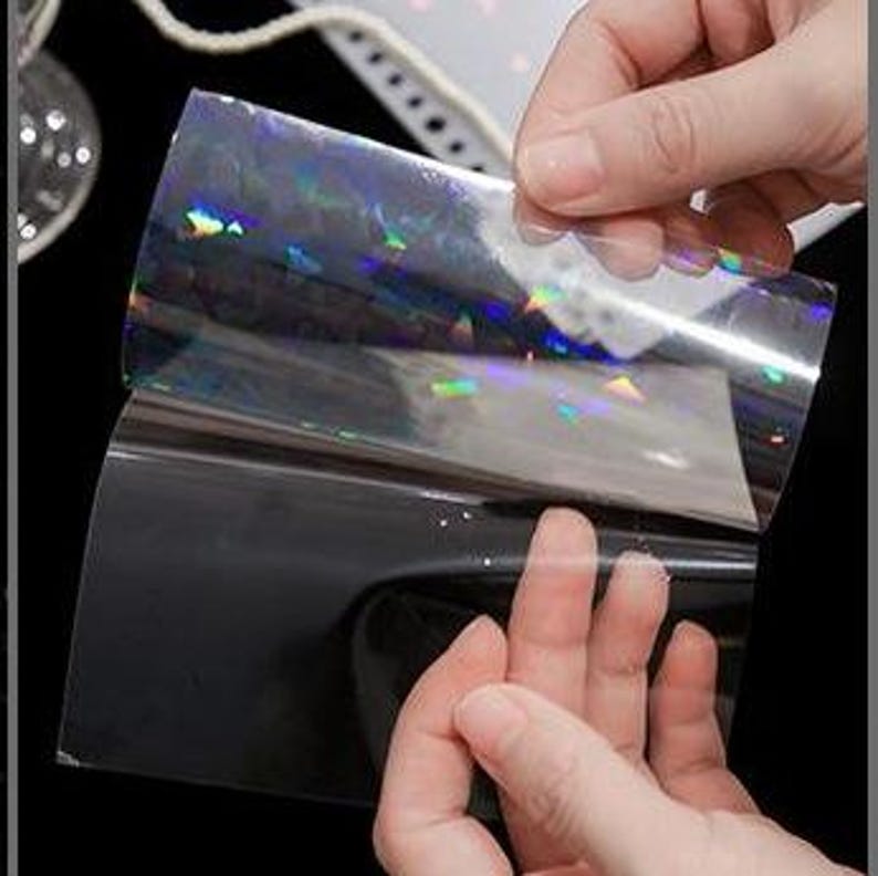 May include: A rectangular sheet of transparent adhesive film featuring a holographic finish that reflects rainbow colors. The film is partially peeled away from its solid black backing, revealing the reflective surface and the clear material.