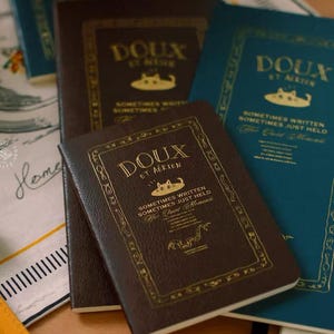 May include: Three rectangular notebooks in dark brown and teal with gold foil lettering. The covers feature the text "DOUX ET AÉRIEN", a cat illustration, and the phrase "SOMETIMES WRITTEN SOMETIMES JUST HELD" framed by a decorative border.