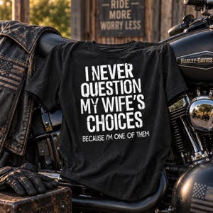 May include: A black t-shirt featuring white distressed text that reads I NEVER QUESTION MY WIFE'S CHOICES BECAUSE I'M ONE OF THEM. The shirt is displayed against a motorcycle and a leather jacket, with a sign in the background reading RIDE MORE WORRY LESS.