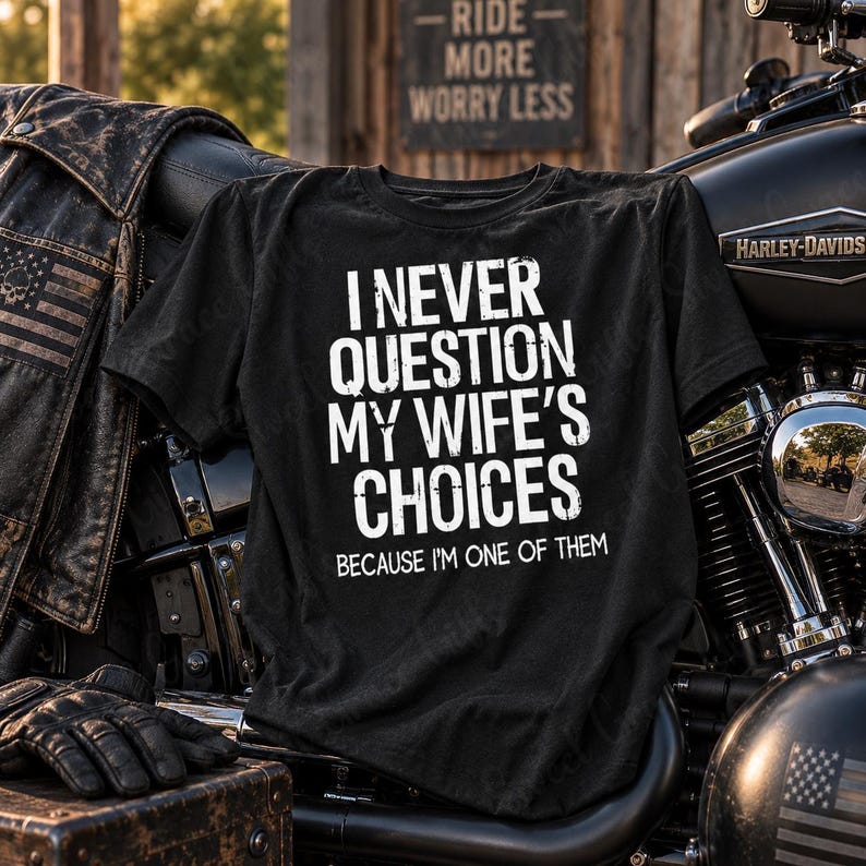 May include: A black t-shirt featuring white distressed text that reads I NEVER QUESTION MY WIFE'S CHOICES BECAUSE I'M ONE OF THEM. The shirt is displayed against a motorcycle and a leather jacket, with a sign in the background reading RIDE MORE WORRY LESS.