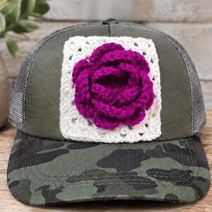 May include: A camouflage print trucker hat featuring a white crocheted square patch on the front panel. A three-dimensional, deep magenta crocheted rose is centered on the white square patch. The hat has a dark green mesh back and a camo brim.