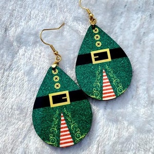 May include: A pair of green glitter Christmas elf-themed dangle earrings with gold hooks. The earrings are shaped like a teardrop and feature a black belt with a gold buckle and a red and white striped Christmas tree.