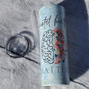 May include: A white tumbler with a blue and white marble design. The tumbler has a black outline of a brain and flowers on one side and the text "Mental Health Matters" on the other side. The tumbler comes with a clear lid and a clear straw.