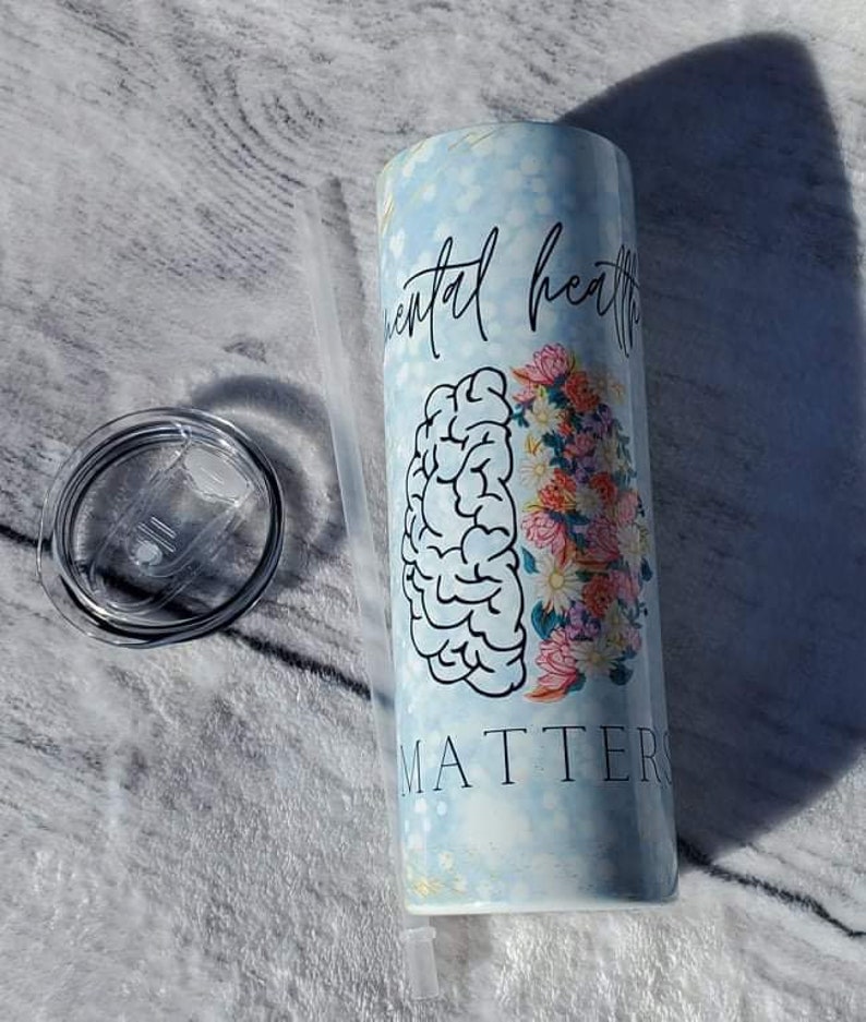 May include: A white tumbler with a blue and white marble design. The tumbler has a black outline of a brain and flowers on one side and the text "Mental Health Matters" on the other side. The tumbler comes with a clear lid and a clear straw.