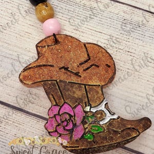 May include: A brown cowboy boot-shaped car air freshener with a pink flower and green leaves. The air freshener is attached to a string of black, tan, and pink beads. The text "Sweet Grace Gifts" is visible.