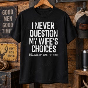 May include: A black short-sleeve t-shirt featuring white distressed text that reads I NEVER QUESTION MY WIFE'S CHOICES BECAUSE I'M ONE OF THEM. The shirt is displayed on a wooden hanger against a rustic background with various decorative items.
