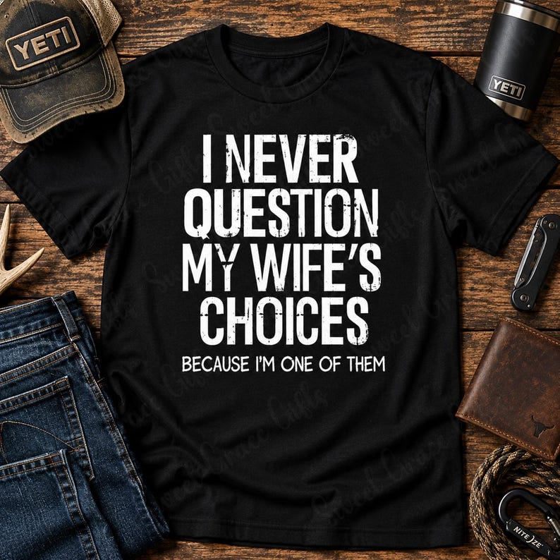 May include: A black t-shirt featuring white distressed text that reads I NEVER QUESTION MY WIFE'S CHOICES BECAUSE I'M ONE OF THEM. The shirt has a crew neckline and short sleeves, presented as a flat lay garment with a simple, bold graphic design.
