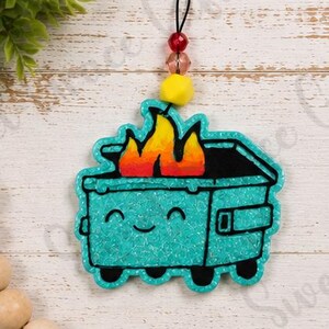May include: A teal dumpster ornament with a smiling face and black outlines, featuring bright orange and yellow flames emerging from the top. The ornament hangs from a black cord adorned with a red bead, a translucent pink bead, and a yellow bead.